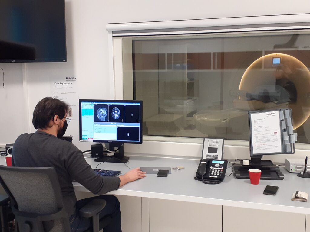 A man with a mouth mask is sitting behind a computer watching at MRI scanning images. Behind the computer is a window and there after a MRI scanner.